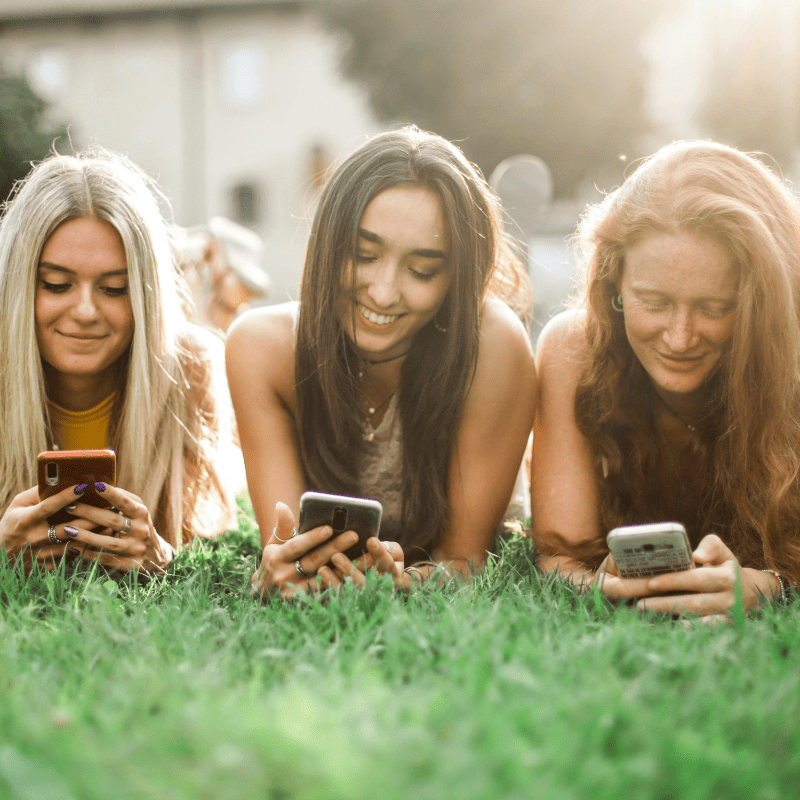 tres garotas da geracao z deitadas na grama mexendo cada uma no seu celular
